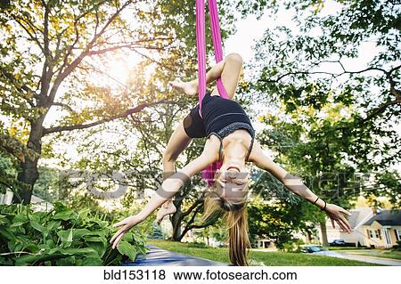 Stock Photo - Acrobatic Caucasian girl hanging on fabric under tree. Fotosearch