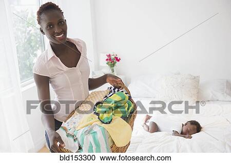 Stock Image - Black mother carrying laundry near sleeping baby boy in bedroom. Fotosearch
