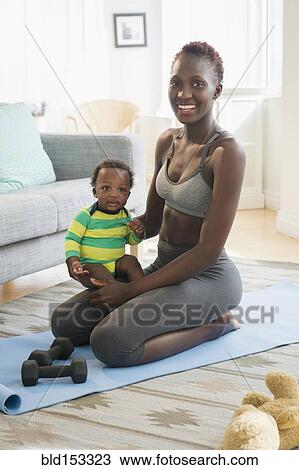 Stock Image - Black mother holding baby boy on exercise mat in living room. Fotosearch