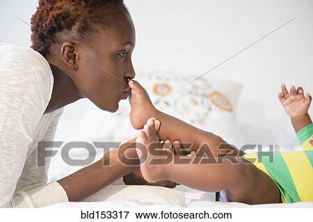 Stock Photo - Black mother kissing feet of baby boy on bed. Fotosearch