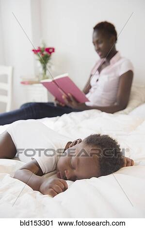 Stock Image - Black mother reading near sleeping baby boy. Fotosearch