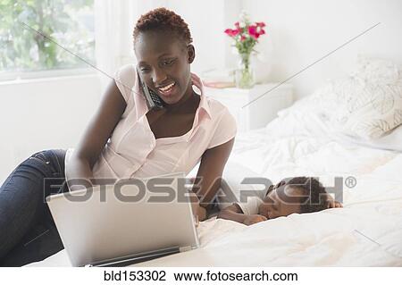 Stock Image - Black mother using laptop near sleeping baby boy. Fotosearch