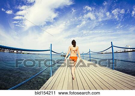 Stock Image - Caucasian girl walking on wooden dock over sea. Fotosearch