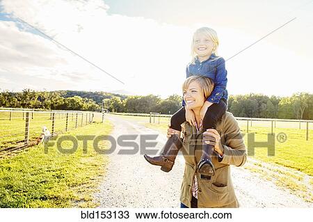 Stock Image - Caucasian mother carrying daughter on shoulders on rural road. Fotosearch