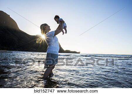 Stock Photo - Caucasian mother holding baby in ocean on beach. Fotosearch