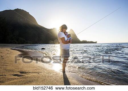 Stock Photograph - Caucasian mother holding baby in waves on beach. Fotosearch