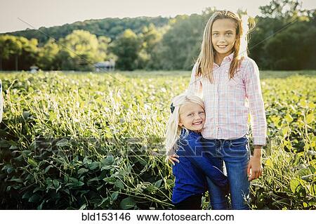 Stock Photograph - Caucasian sisters hugging in crop field on farm. Fotosearch
