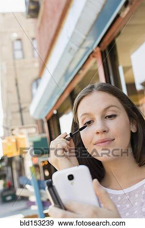 Stock Photo - Caucasian woman applying makeup in cell phone camera. Fotosearch