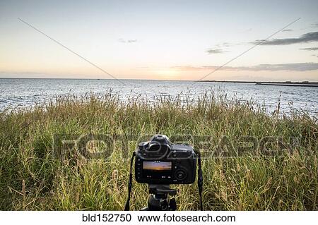 Stock Image - Close up of camera photographing sunset over ocean horizon. Fotosearch