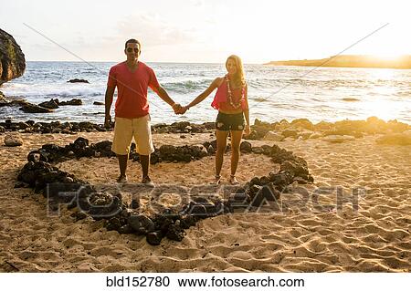 Stock Image - Couple holding hands in heart shape on beach. Fotosearch