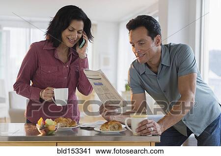 Stock Image - Couple reading newspaper and talking on cell phone at breakfast. Fotosearch