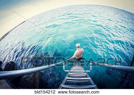 Stock Image - Fish-eye lens view of older Caucasian man descending ladder into ocean. Fotosearch