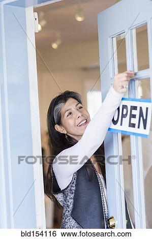 Stock Photograph - Hispanic small business owner hanging open sign on door. Fotosearch