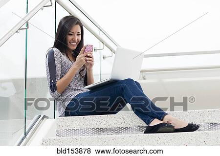 Stock Photo - Hispanic woman using cell phone and laptop on staircase. Fotosearch