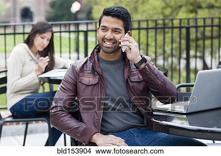Picture - Indian man talking on cell phone at table outdoors. Fotosearch