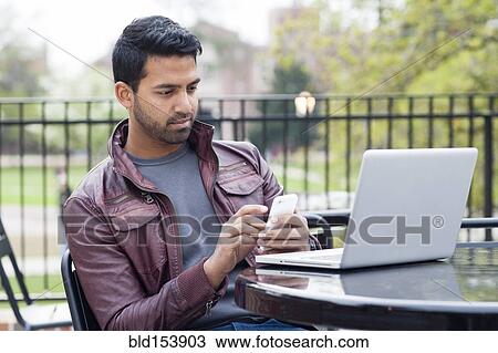 Indian man using cell phone and laptop at table outdoors View Large Photo Image Stock Image - Indian man using cell phone and laptop at table outdoors. Fotosearch