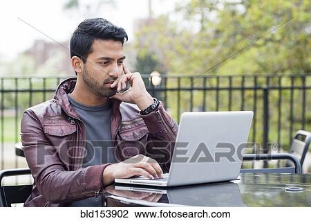 Stock Image - Indian man using laptop at table outdoors. Fotosearch