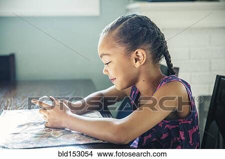 Picture - Mixed race girl using cell phone at kitchen table. Fotosearch
