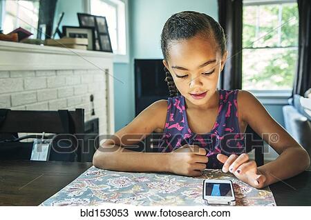 Stock Image - Mixed race girl using cell phone at kitchen table. Fotosearch