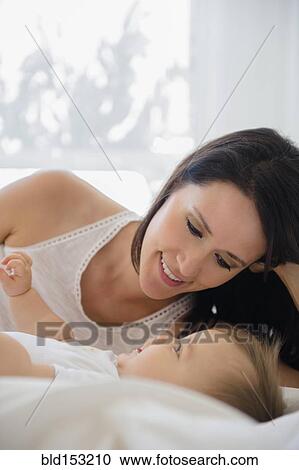 Stock Image - Mixed race mother laying with baby on bed. Fotosearch