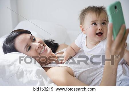 Stock Photo - Mixed race mother playing with baby and cell phone on bed. Fotosearch