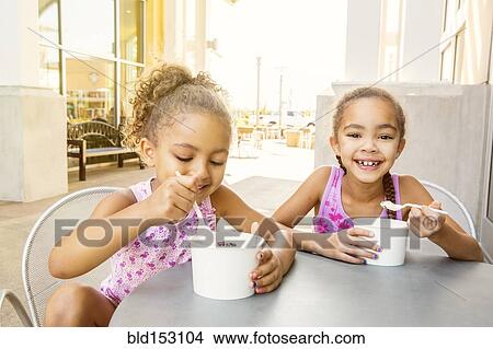 Picture - Mixed race sisters eating ice cream at outdoor cafe. Fotosearch