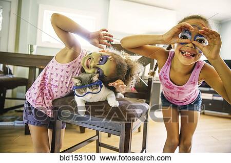 Stock Image - Mixed race sisters playing with face paint and googly eyes. Fotosearch