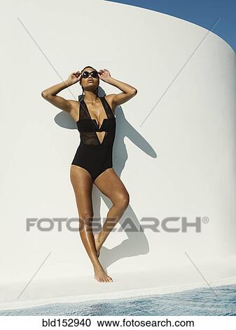 Stock Image - Mixed race woman in swimsuit wearing sunglasses near swimming pool. Fotosearch