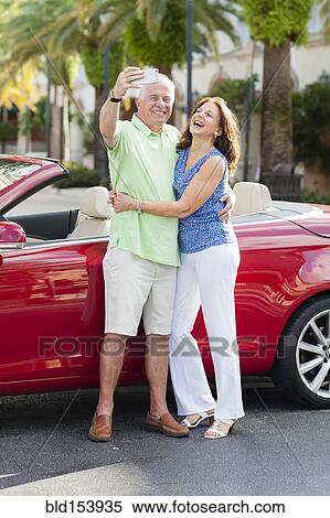 Stock Photography - Older Caucasian couple taking cell phone photograph near convertible. Fotosearch
