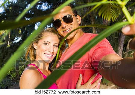 Stock Image - Smiling couple hugging behind palm frond leaves. Fotosearch