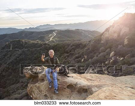 Stock Image - Older Caucasian man taking cell phone photograph on rocky hilltop. Fotosearch
