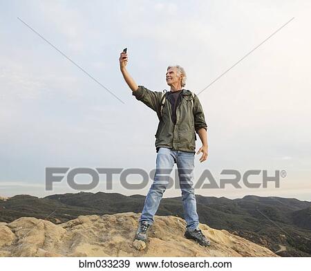 Stock Photo - Older Caucasian man taking cell phone photograph on rocky hilltop. Fotosearch