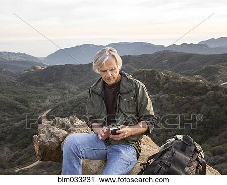 Stock Image - Older Caucasian man using cell phone on rocky hilltop. Fotosearch