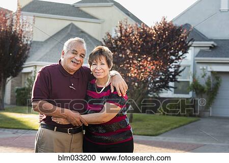 Stock Image - Older Hispanic couple smiling outside suburban home. Fotosearch