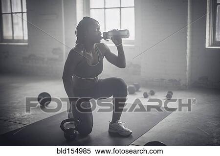 Stock Photography - Black woman drinking water bottle in dark gym. Fotosearch