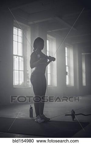Stock Image - Black woman lifting weights in dark gym. Fotosearch