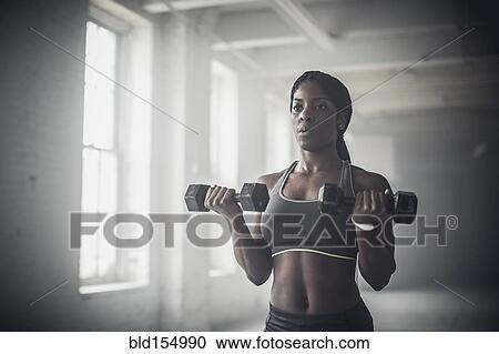 Stock Image - Black woman lifting weights in dark gym. Fotosearch