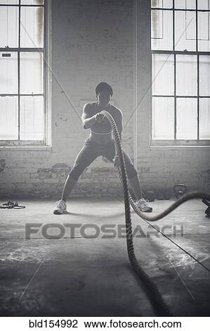 Stock Image - Black woman tossing ropes in dark gym. Fotosearch