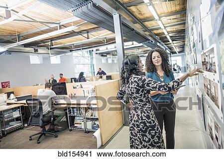 Stock Image - Businesswomen examining photographs on wall in office. Fotosearch