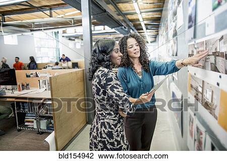 Stock Image - Businesswomen examining photographs on wall in office. Fotosearch