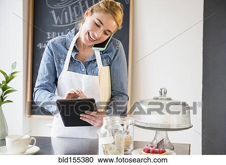 Stock Image - Caucasian barista using cell phone and digital tablet in cafe. Fotosearch