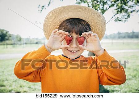 Stock Photo - Caucasian boy holding strawberries over eyes on farm. Fotosearch
