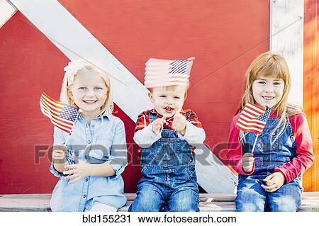 Stock Image - Caucasian children waving American flags outside barn. Fotosearch