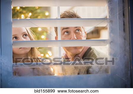 Stock Photo - Caucasian father and daughter peering out frosty window at Christmas. Fotosearch