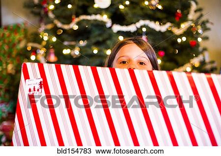 Stock Image - Caucasian girl holding wrapped Christmas gift near tree. Fotosearch