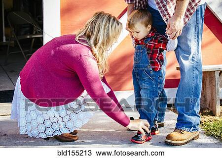 Stock Image - Caucasian mother tying shoes of son outside barn. Fotosearch