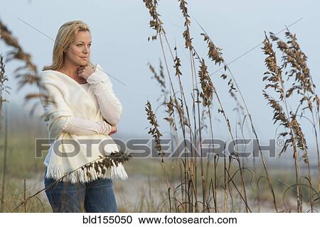 Stock Image - Caucasian woman standing in tall grass on beach. Fotosearch