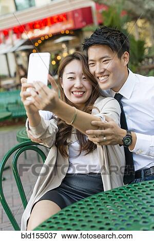 Stock Photo - Korean couple taking cell phone photograph at sidewalk cafe. Fotosearch