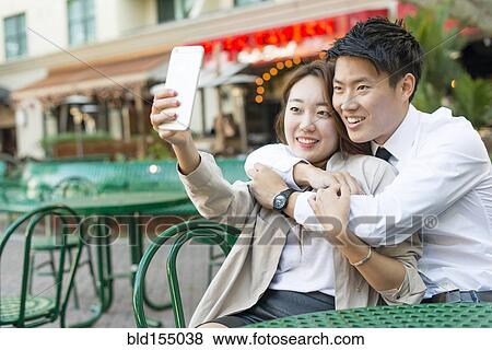 Stock Photo - Korean couple taking cell phone photograph at sidewalk cafe. Fotosearch