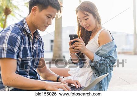 Stock Photograph - Korean couple using laptop and cell phone outdoors. Fotosearch